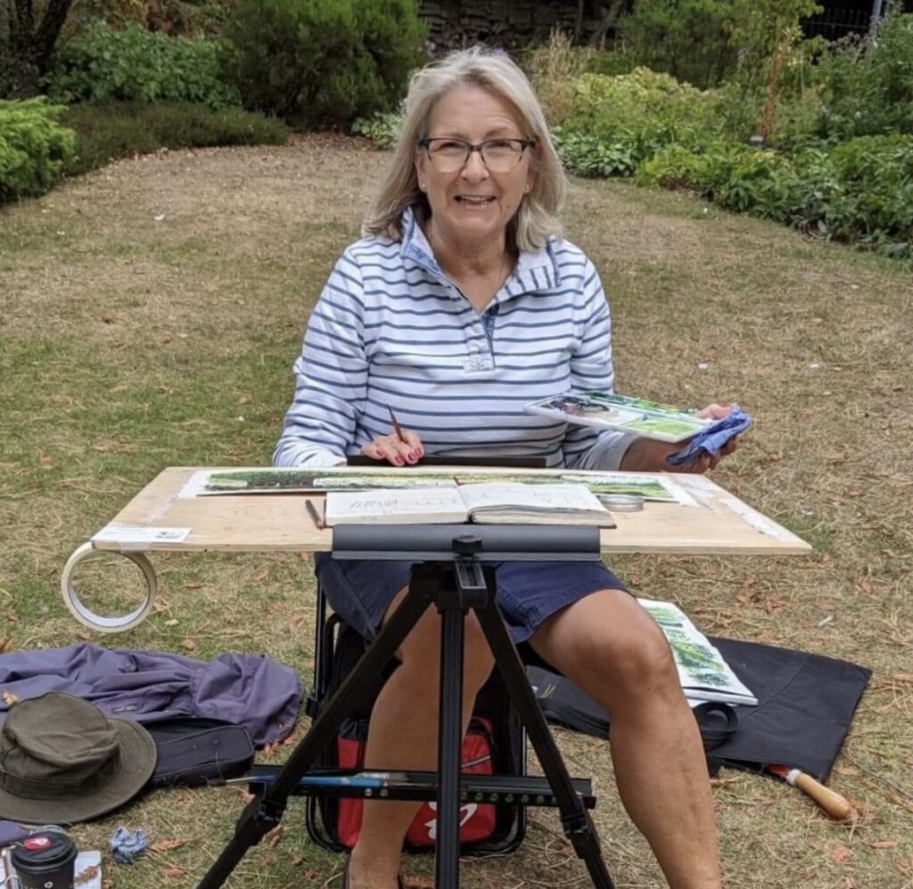 Artist pictured smiling at the camera, sitting on a stool working at an outdoor easel.