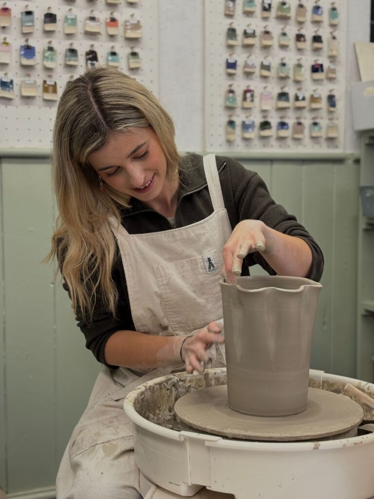 A person is seated at a pottery wheel in a light-filled studio, carefully throwing a large vase from a mound of spinning clay. Their hands are steady and focused, shaping the clay as it rises upward into a tall, symmetrical form. Water glistens on the surface as the vessel takes shape beneath their fingers. The studio around them is calm and creative, with pottery tools, trimmed clay scraps, and finished pieces nearby.