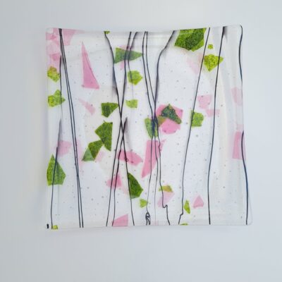 A large, square fused glass plate.  The glass is mainly clear with pink and green shapes and thin black lines across it.